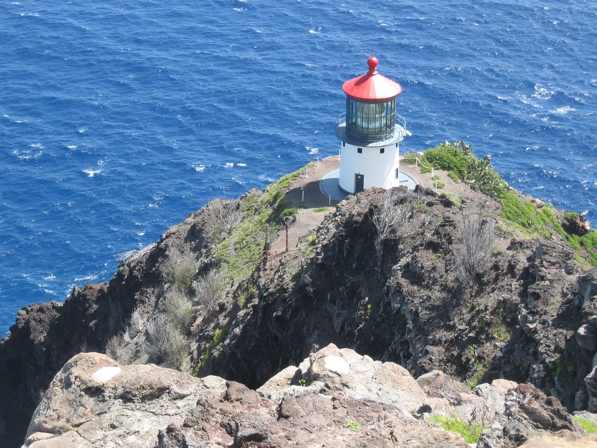 Makapuʻu Lighthouse on the rocky cliff below the hiking trail, above blue ocean.